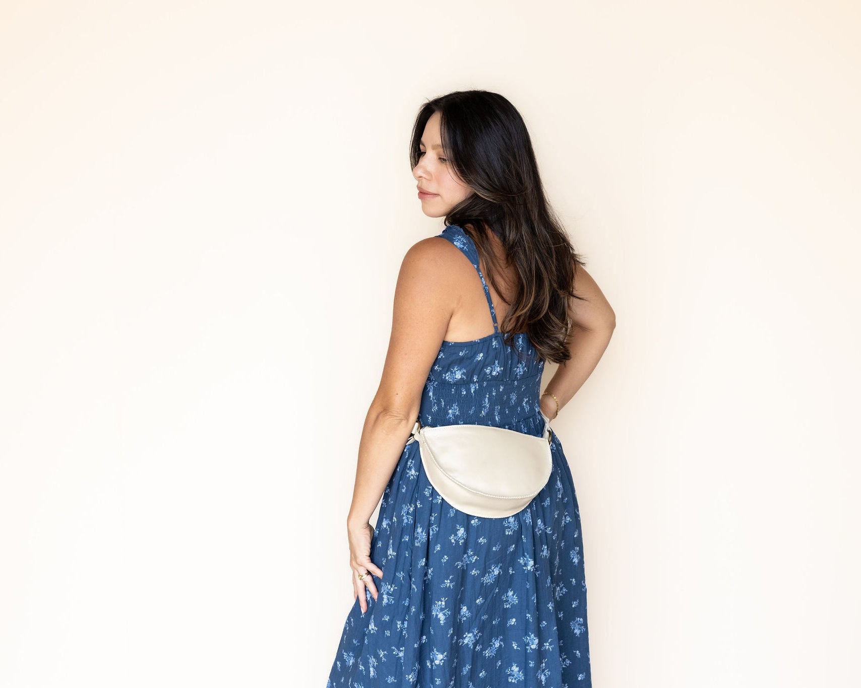 A woman wearing a blue floral print dress and a cream-colored crescent-shaped crossbody bag, standing against a plain off-white wall.