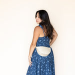 A woman wearing a blue floral print dress and a cream-colored crescent-shaped crossbody bag, standing against a plain off-white wall.