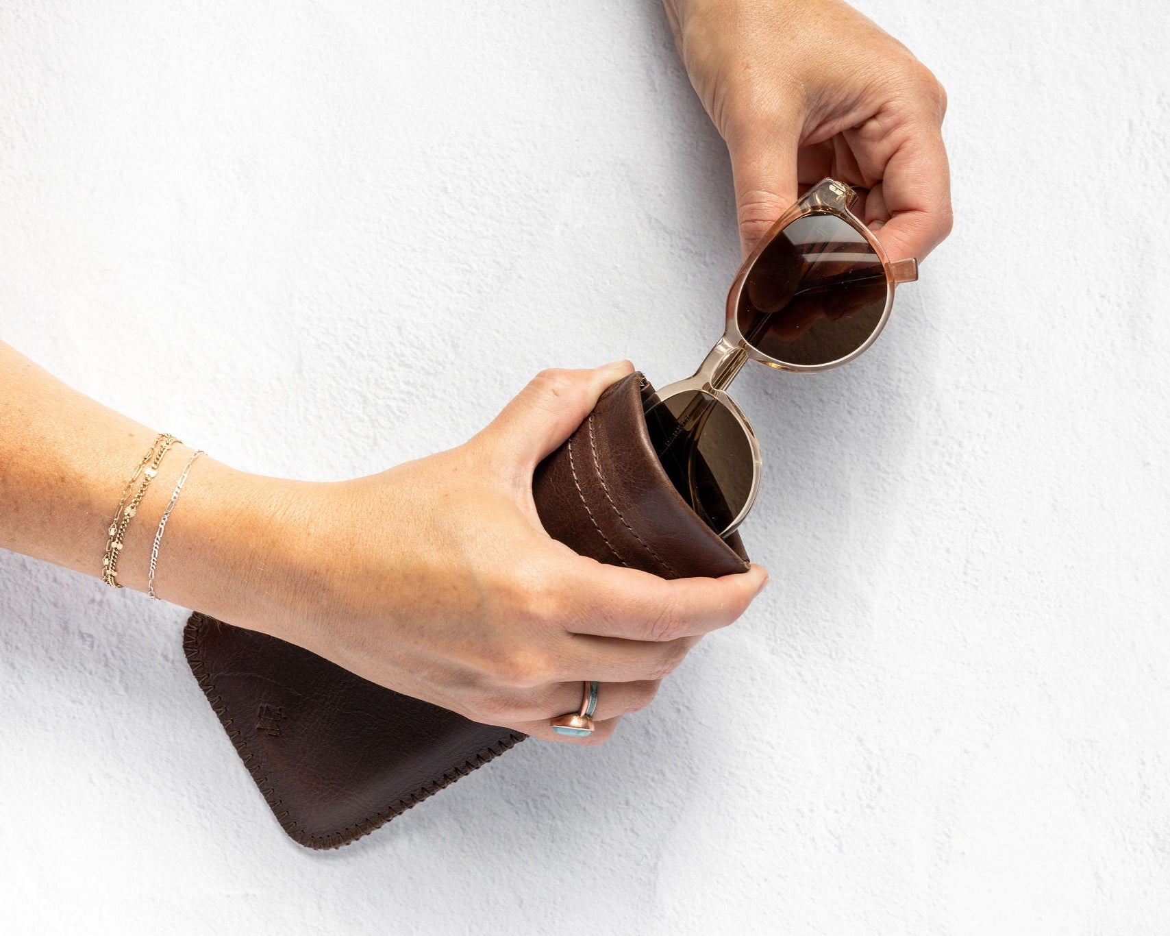 A person's hands holding a dark brown leather sunglasses case while inserting a pair of round sunglasses with dark lenses. The person is wearing a delicate gold chain bracelet on one wrist, set against a clean, textured white background.