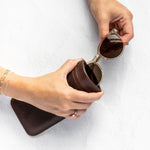 A person's hands holding a dark brown leather sunglasses case while inserting a pair of round sunglasses with dark lenses. The person is wearing a delicate gold chain bracelet on one wrist, set against a clean, textured white background.