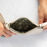 A cream-colored leather wristlet clutch shown open, held by two hands against a textured white background. The clutch features a dark green patterned interior lining and a cream strap.