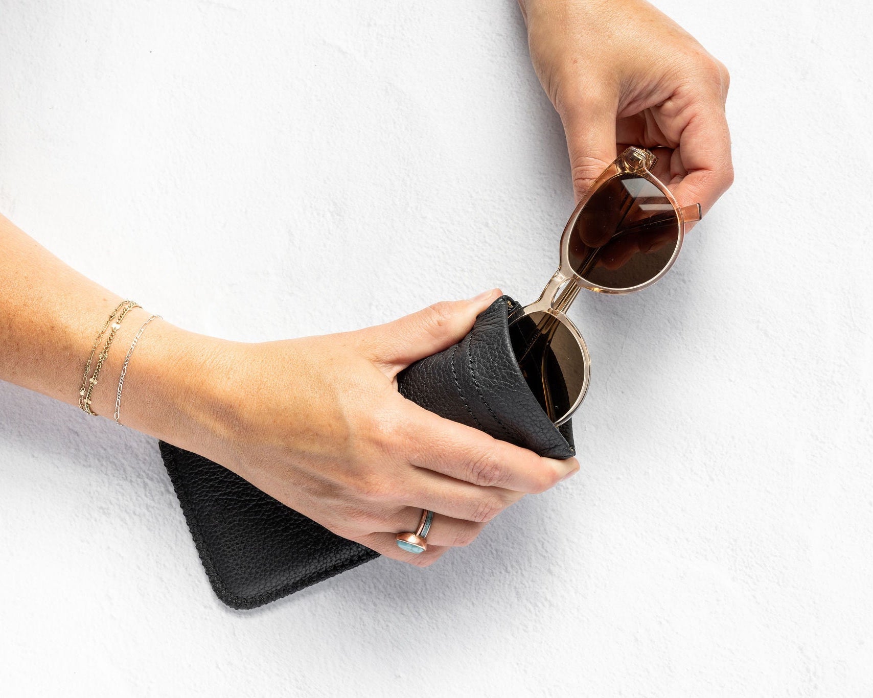 A person's hands holding a black textured leather pouch and a pair of round sunglasses with gold frames and dark lenses, set against a clean white textured background.