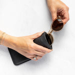 A person's hands holding a black textured leather pouch and a pair of round sunglasses with gold frames and dark lenses, set against a clean white textured background.