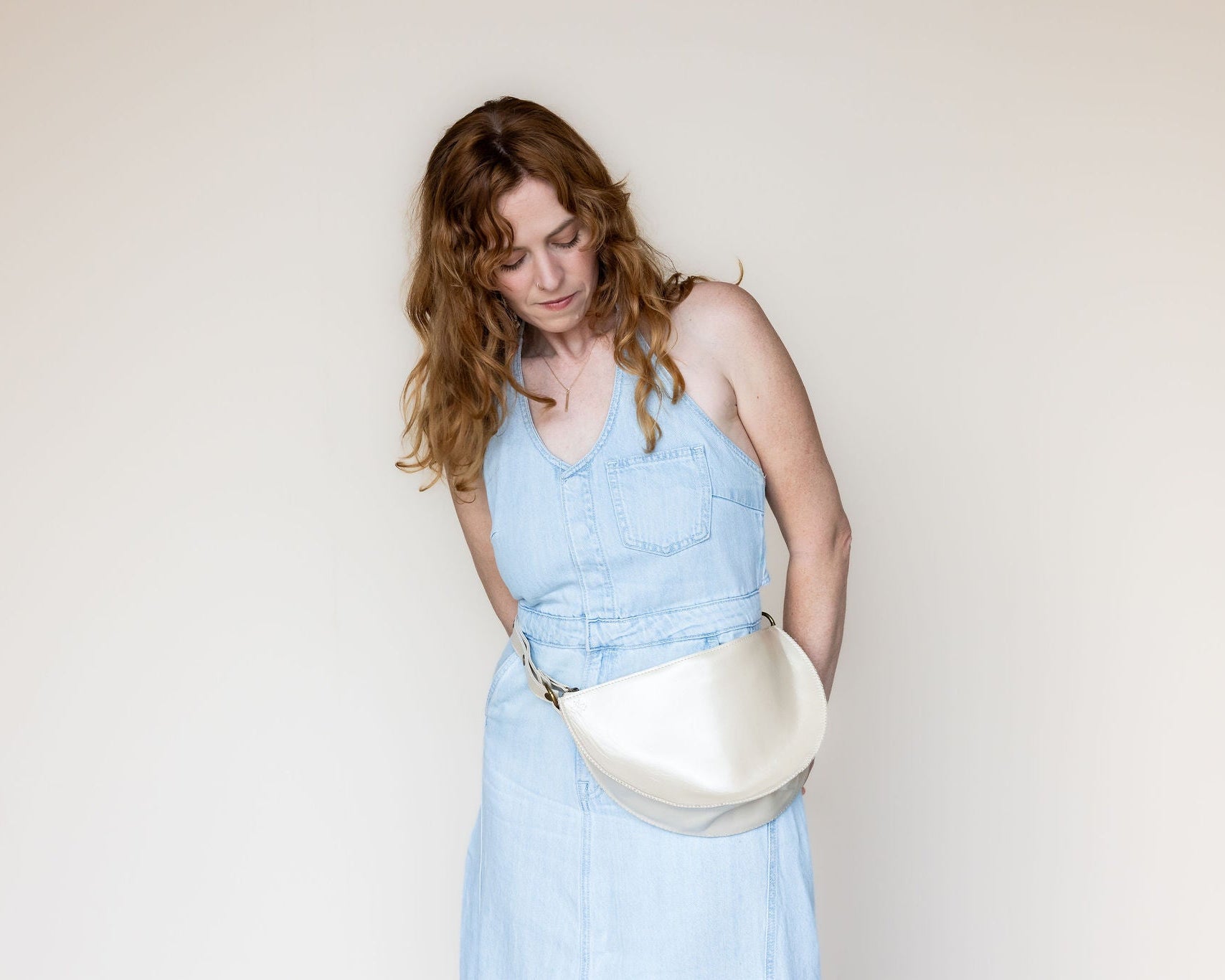A woman with wavy light brown hair wearing a light blue denim dress and a cream-colored crescent-shaped leather belt bag, standing against a plain off-white background.