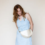 A woman with wavy light brown hair wearing a light blue denim dress and a cream-colored crescent-shaped leather belt bag, standing against a plain off-white background.