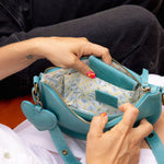 A close-up of a person holding open a small turquoise leather shoulder bag, showing the patterned interior lining and zipper closure.