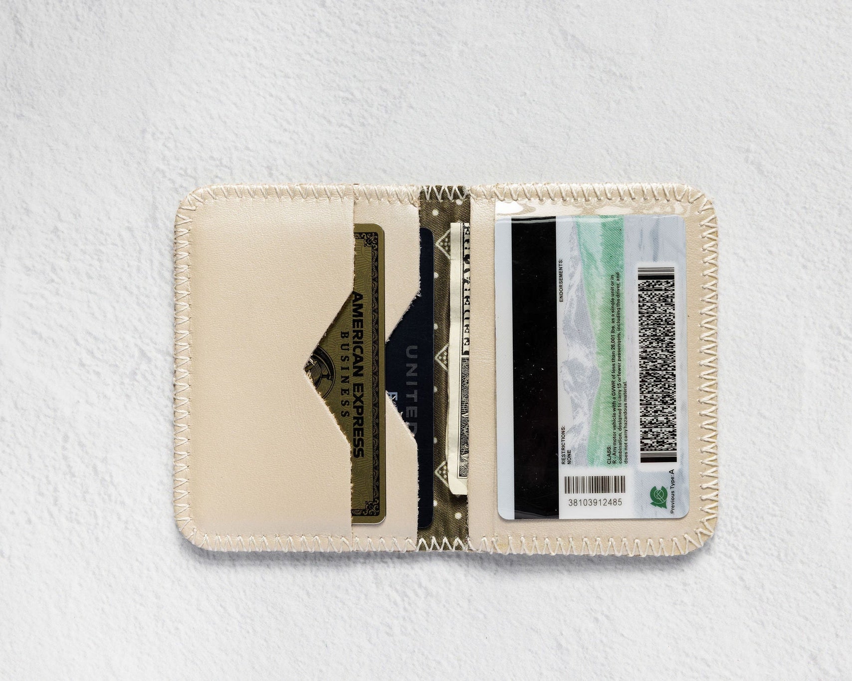 A cream-colored leather card holder wallet shown open, displaying its interior slots filled with cards and cash, set against a textured white background.
