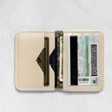 A cream-colored leather card holder wallet shown open, displaying its interior slots filled with cards and cash, set against a textured white background.