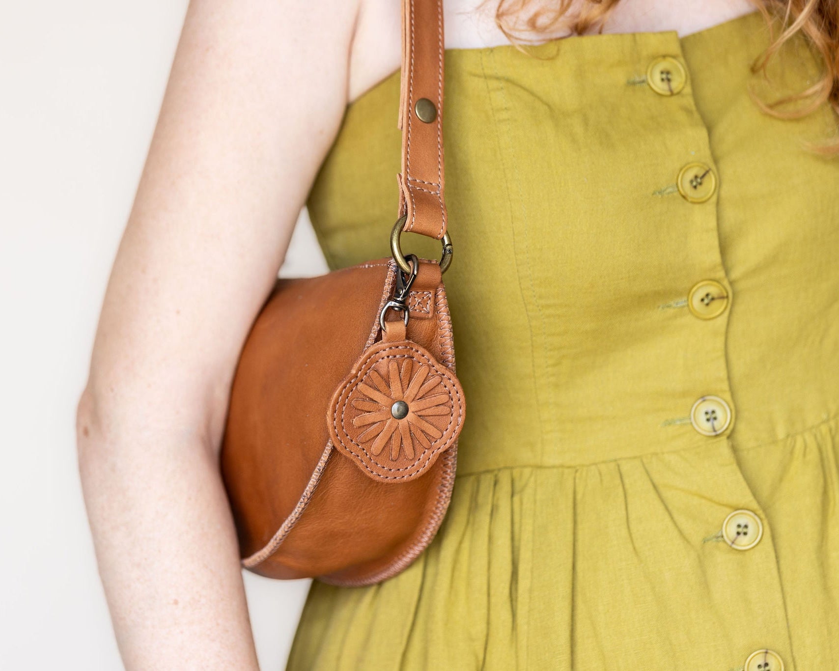 A close-up of a person wearing a mustard yellow dress and carrying a brown leather shoulder bag with a decorative flower-shaped leather charm.