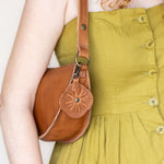 A close-up of a person wearing a mustard yellow dress and carrying a brown leather shoulder bag with a decorative flower-shaped leather charm.