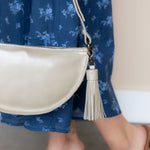 A cream-colored leather half-moon crossbody bag with a tassel charm, worn by a person in a blue floral dress.