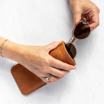 A person's hands holding a pair of sunglasses over a brown leather case on a white textured background.
