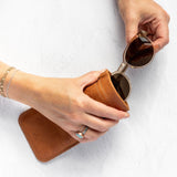 A person's hands holding a pair of sunglasses over a brown leather case on a white textured background.