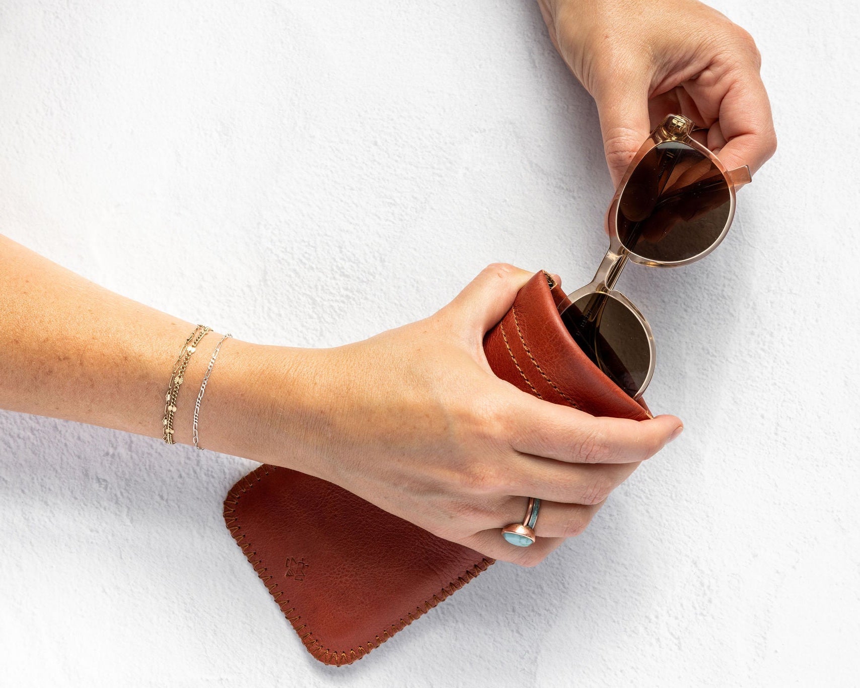 A person's hands holding a pair of round sunglasses over a brown leather case against a white textured background.