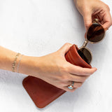 A person's hands holding a pair of round sunglasses over a brown leather case against a white textured background.