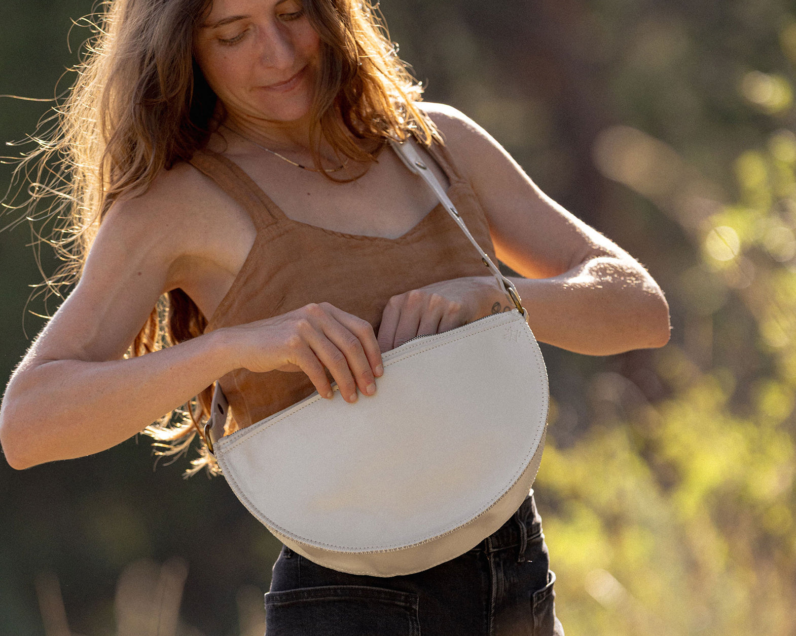 A woman outdoors wearing a brown camisole and dark jeans, holding a cream-colored crescent-shaped shoulder bag against a blurred natural background.