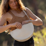 A woman outdoors wearing a brown camisole and dark jeans, holding a cream-colored crescent-shaped shoulder bag against a blurred natural background.
