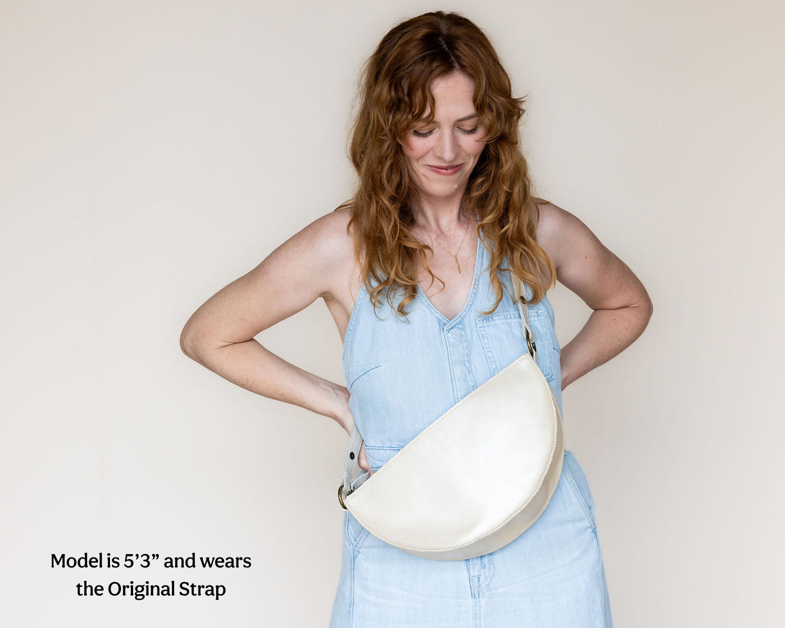 A woman wearing a light blue denim dress models a cream-colored crescent-shaped shoulder bag. The image features a text overlay in the bottom-left corner reading 'Model is 5'3" and wears the Original Strap'.