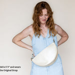 A woman wearing a light blue denim dress models a cream-colored crescent-shaped shoulder bag. The image features a text overlay in the bottom-left corner reading 'Model is 5'3" and wears the Original Strap'.
