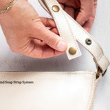 Close-up of a person's hands demonstrating the patented snap strap system on a cream-colored handbag. The strap is shown attaching to the bag with brass snap buttons.