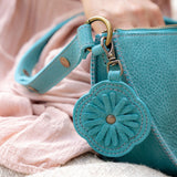 A close-up of a turquoise blue pebbled leather handbag with a matching flower-shaped charm and gold-tone hardware, held by a person wearing a pink dress.