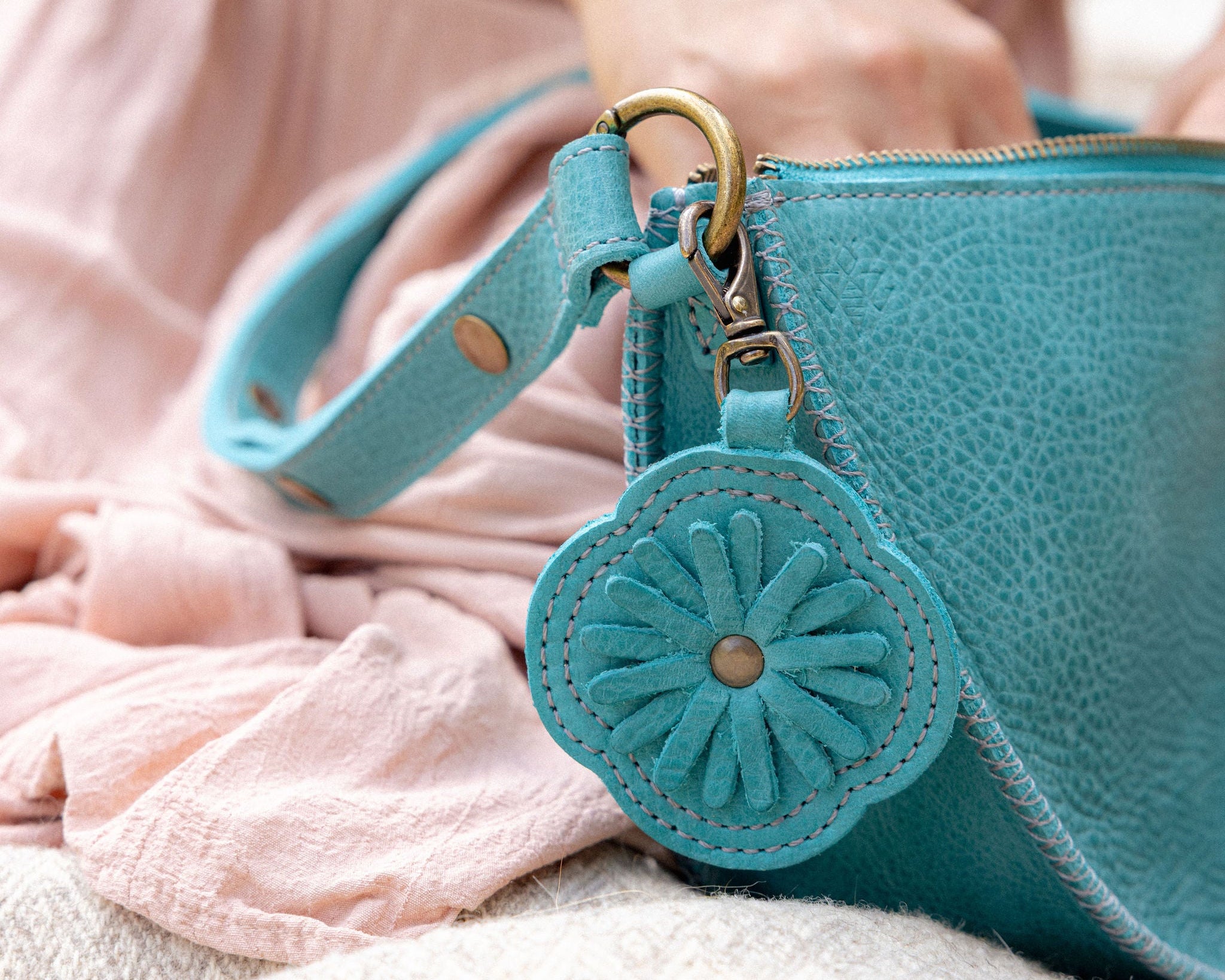 A close-up of a turquoise blue pebbled leather handbag with a matching flower-shaped charm and gold-tone hardware, held by a person wearing a pink dress.