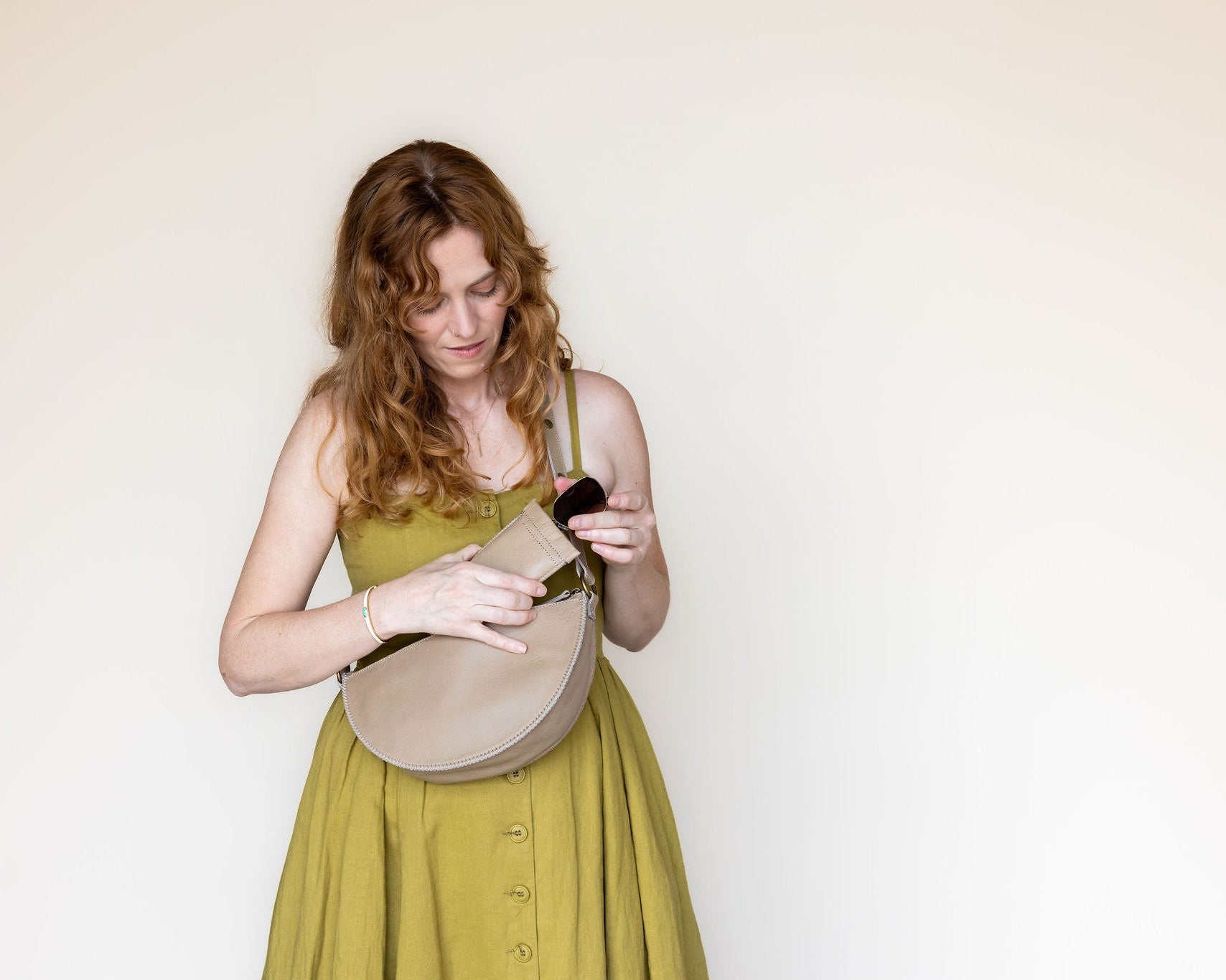 A woman with red hair wearing a yellow-green dress and holding a beige crescent-shaped shoulder bag, looking down inside the bag against a plain off-white background.