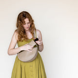 A woman with red hair wearing a yellow-green dress and holding a beige crescent-shaped shoulder bag, looking down inside the bag against a plain off-white background.