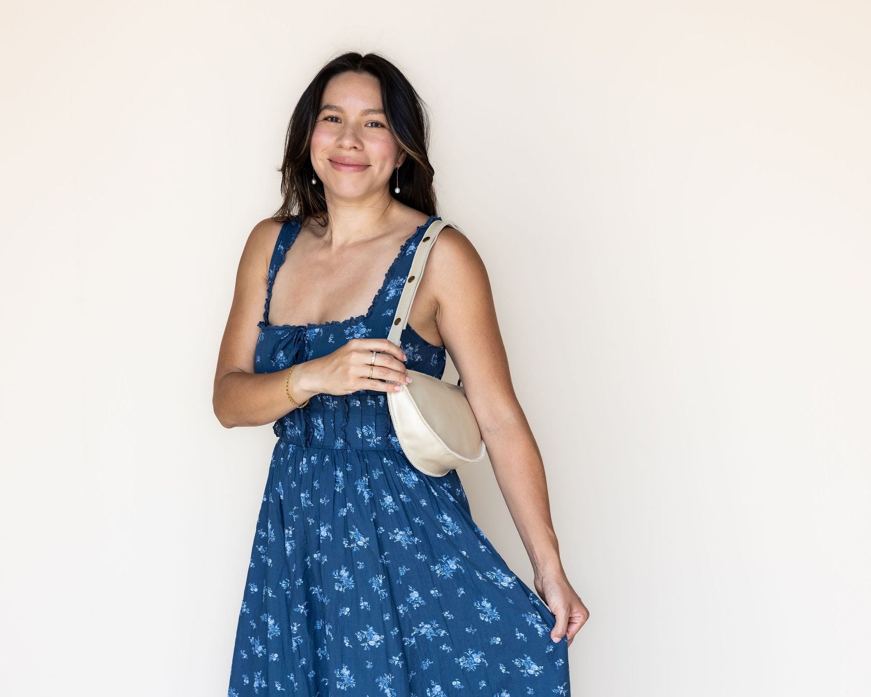 A woman wearing a blue floral print sundress and carrying a cream-colored shoulder bag, standing against a plain off-white wall.