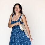 A woman wearing a blue floral print sundress and carrying a cream-colored shoulder bag, standing against a plain off-white wall.