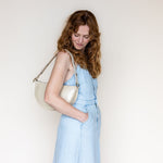 A woman with wavy light brown hair wearing a light blue sleeveless dress and carrying a cream-colored leather shoulder bag, standing against a plain off-white background.
