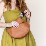 A woman wearing a mustard green dress holds a tan leather crescent shoulder bag and sunglasses against a plain off-white background.