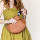 A woman wearing a mustard green dress holds a tan leather crescent shoulder bag and sunglasses against a plain off-white background.