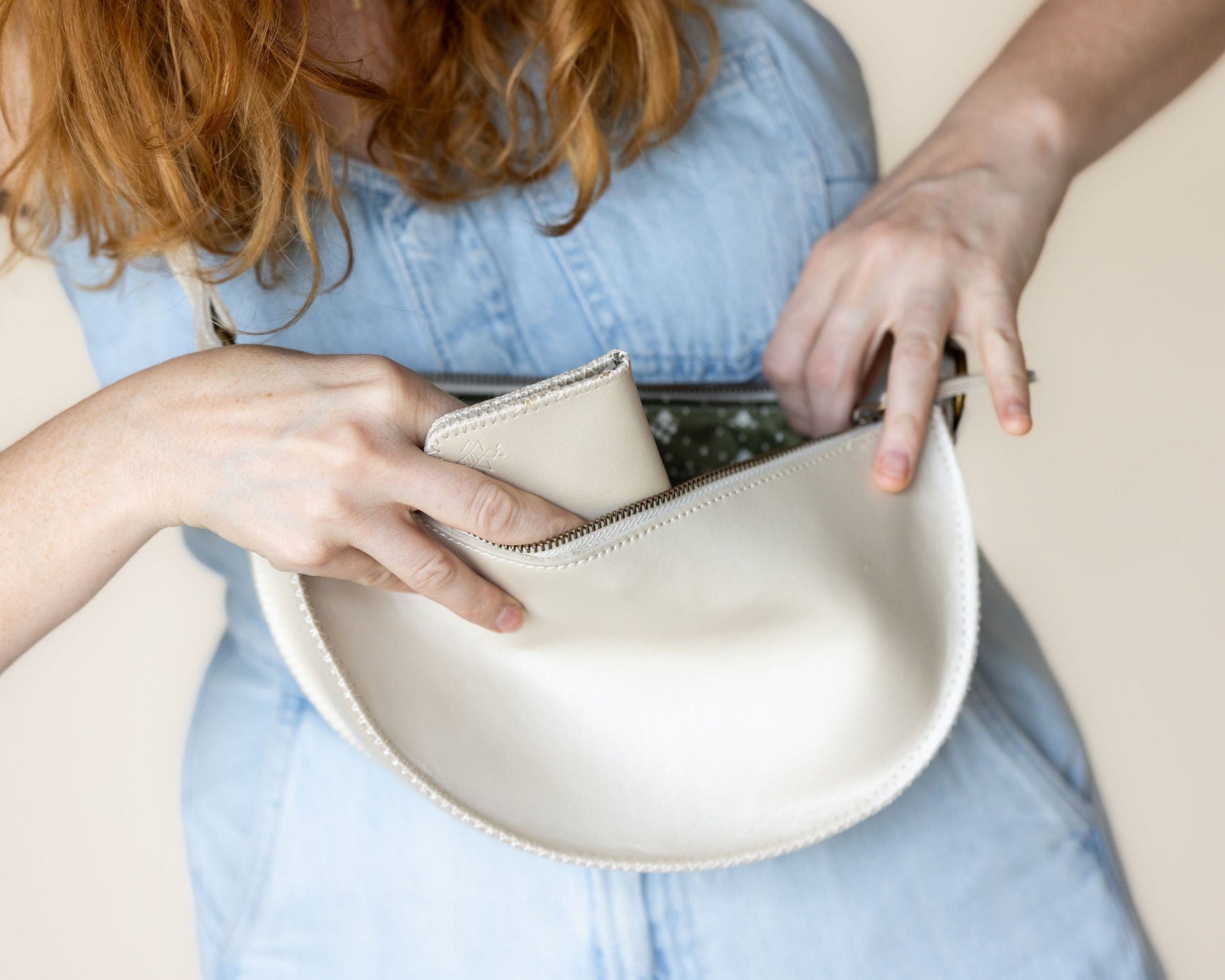 A close-up of a person wearing a light blue denim dress, showing hands placing a cream-colored leather pouch or wallet into a matching curved leather bag.