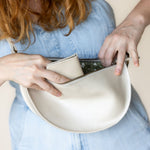 A close-up of a person wearing a light blue denim dress, showing hands placing a cream-colored leather pouch or wallet into a matching curved leather bag.