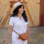 A woman wearing a white short-sleeve button-up dress, a beige wide-brim hat, and a brown leather crossbody bag walks outdoors in front of a wooden barn door while carrying a wicker basket.
