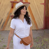 A woman wearing a white short-sleeve button-up dress, a beige wide-brim hat, and a brown leather crossbody bag walks outdoors in front of a wooden barn door while carrying a wicker basket.