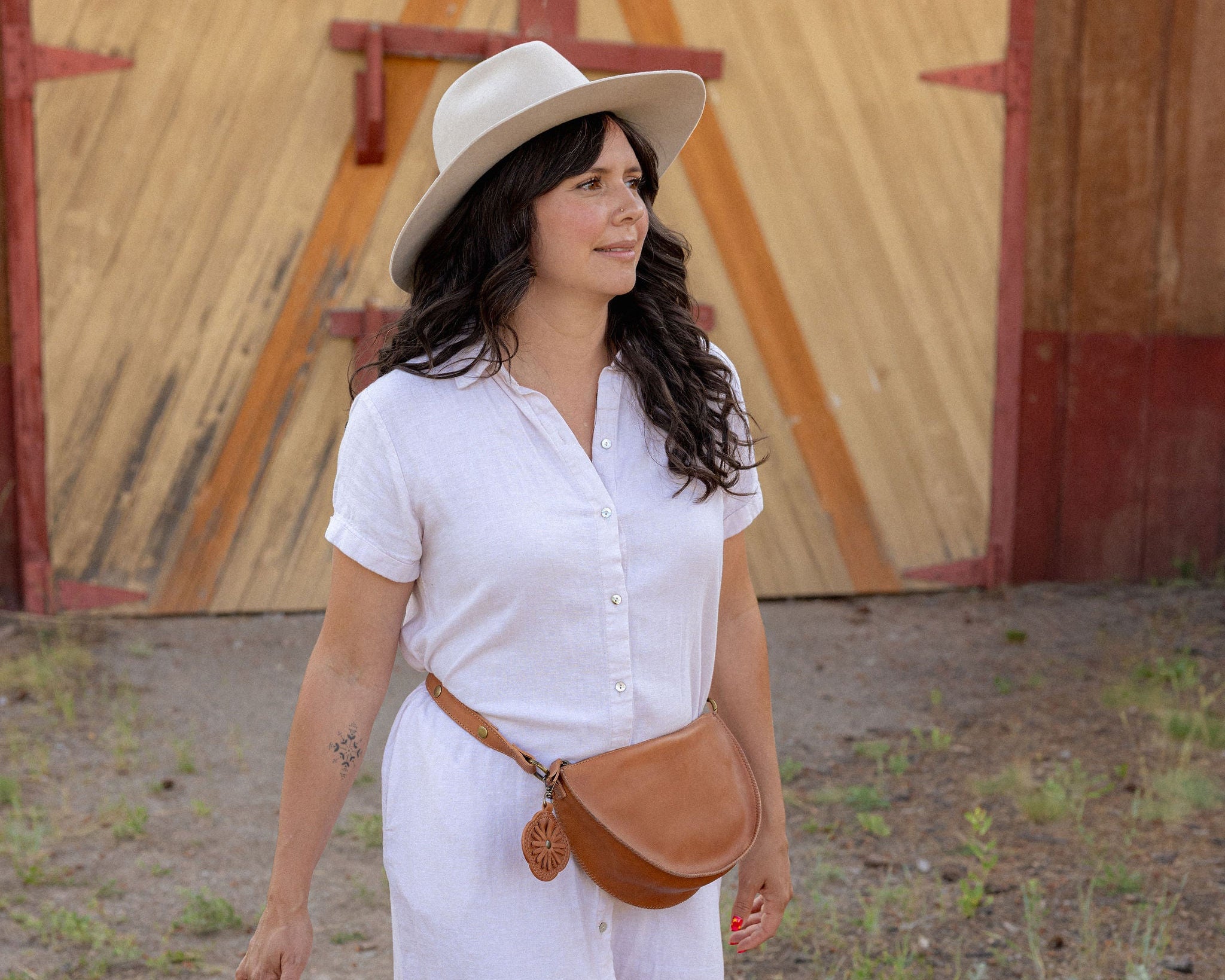 A woman wearing a white short-sleeve button-up dress, a beige wide-brim hat, and a brown leather crossbody bag walks outdoors in front of a wooden barn door while carrying a wicker basket.