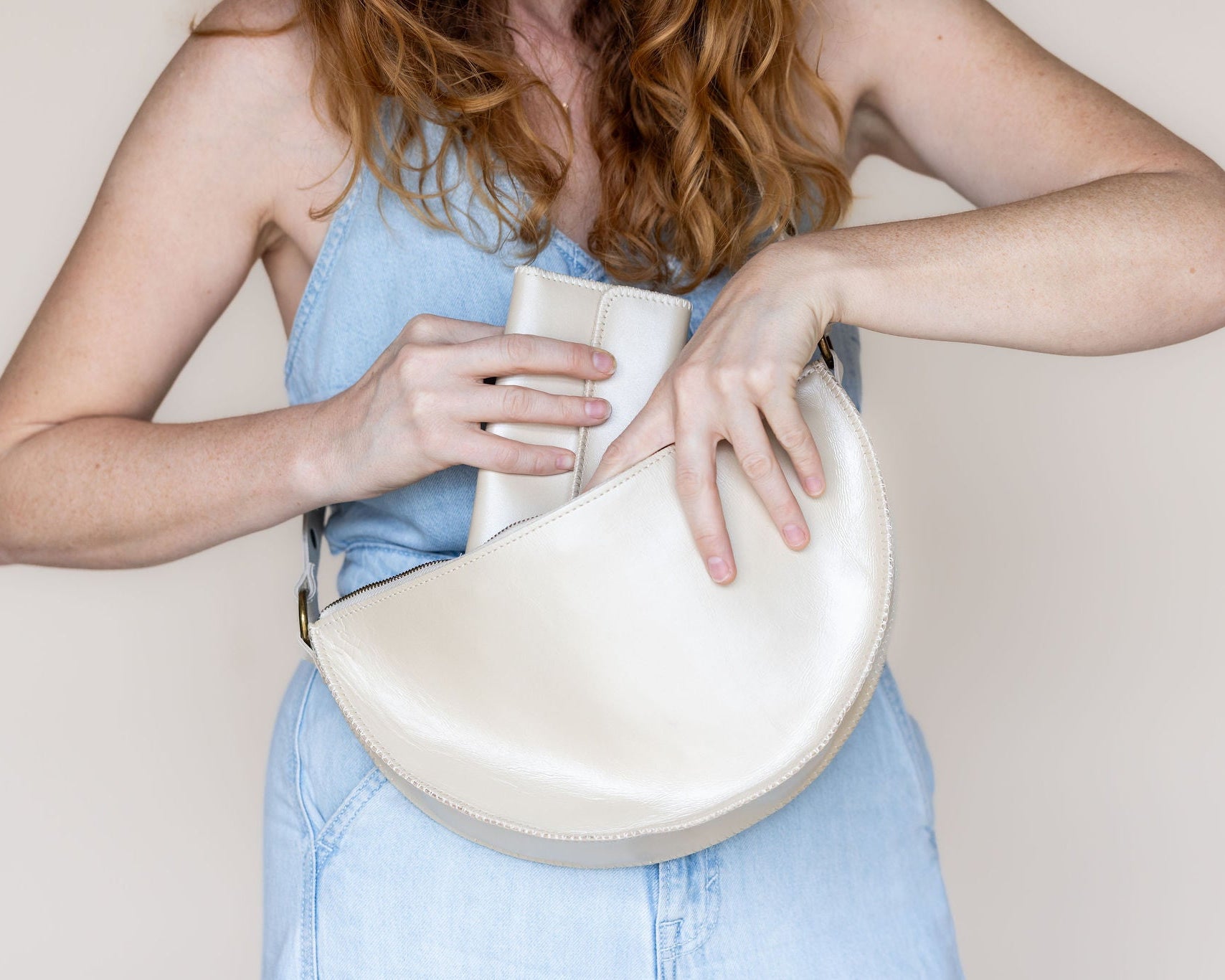 A woman wearing a light blue denim top holds a cream-colored, semi-circular leather shoulder bag against a plain beige background.