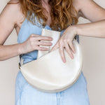 A woman wearing a light blue denim top holds a cream-colored, semi-circular leather shoulder bag against a plain beige background.