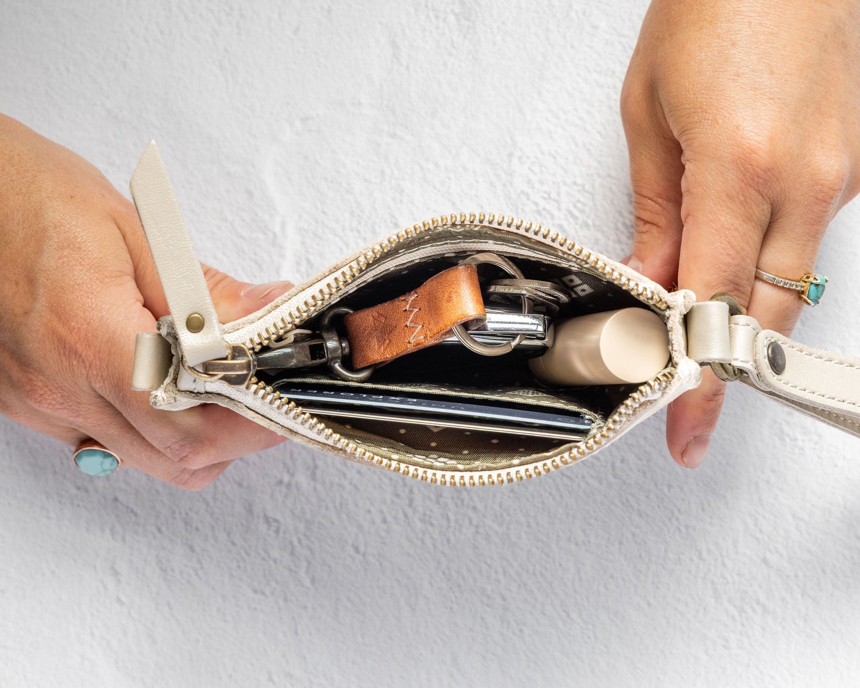 A close-up of a person's hands holding open a small cream-colored leather zippered wallet or clutch purse, showing its contents including cards, a brown leather keychain, and a small cylindrical item, set against a light gray textured background.