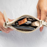 A close-up of a person's hands holding open a small cream-colored leather zippered wallet or clutch purse, showing its contents including cards, a brown leather keychain, and a small cylindrical item, set against a light gray textured background.
