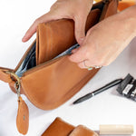 A person's hands are shown opening a tan leather zippered pouch to reveal its interior. A matching tan leather wallet and a blue textured phone case are laid out on a white table alongside other small accessories.