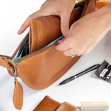 A person's hands are shown opening a tan leather zippered pouch to reveal its interior. A matching tan leather wallet and a blue textured phone case are laid out on a white table alongside other small accessories.