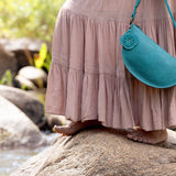 A person wearing a dusty pink tiered maxi dress stands barefoot on a large rock outdoors, holding a teal crescent-shaped shoulder bag with a matching strap.