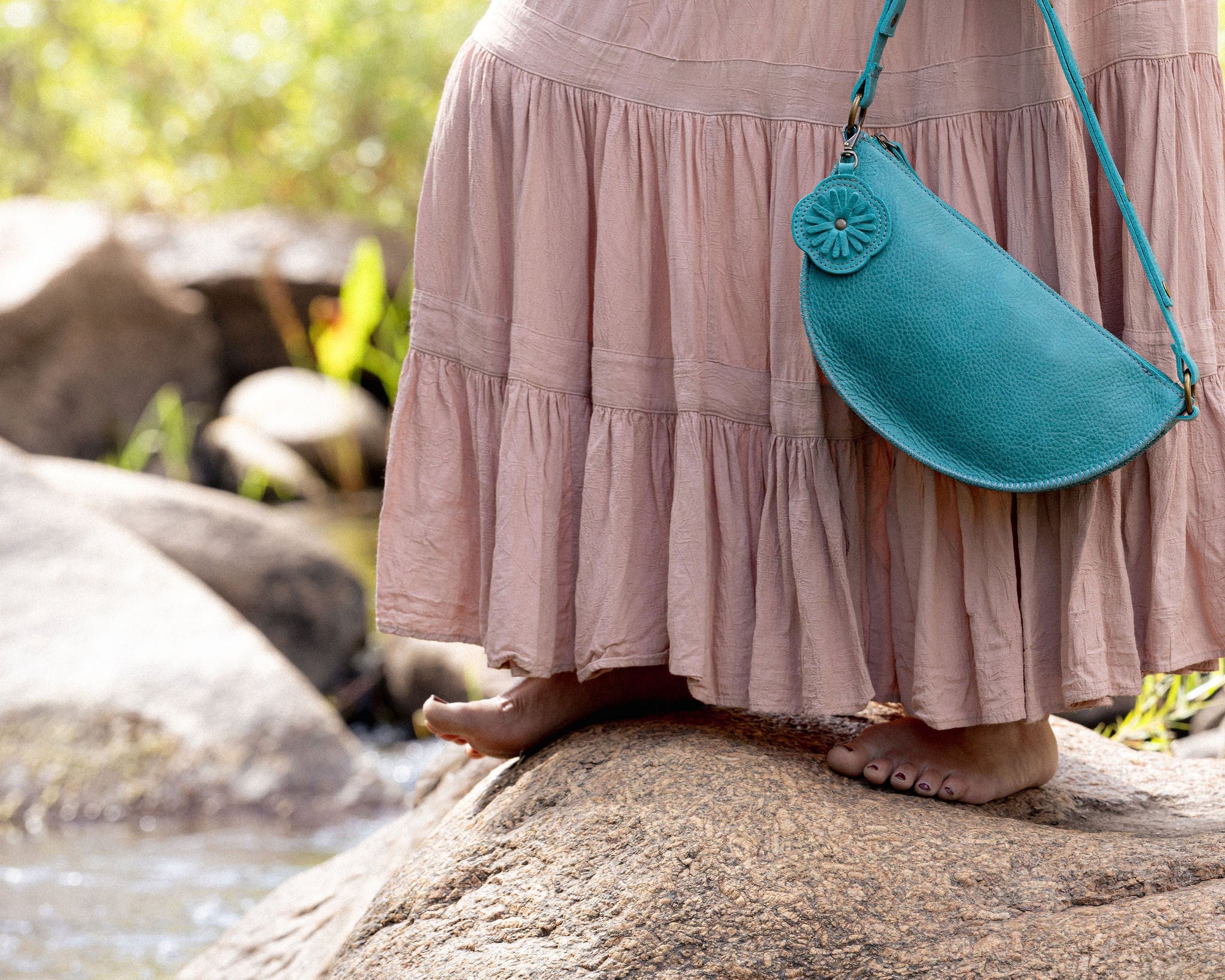 A person wearing a dusty pink tiered maxi dress stands barefoot on a large rock outdoors, holding a teal crescent-shaped shoulder bag with a matching strap.