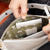 A close-up of a person's hands opening a white leather crossbody bag to show its olive green patterned interior lining.