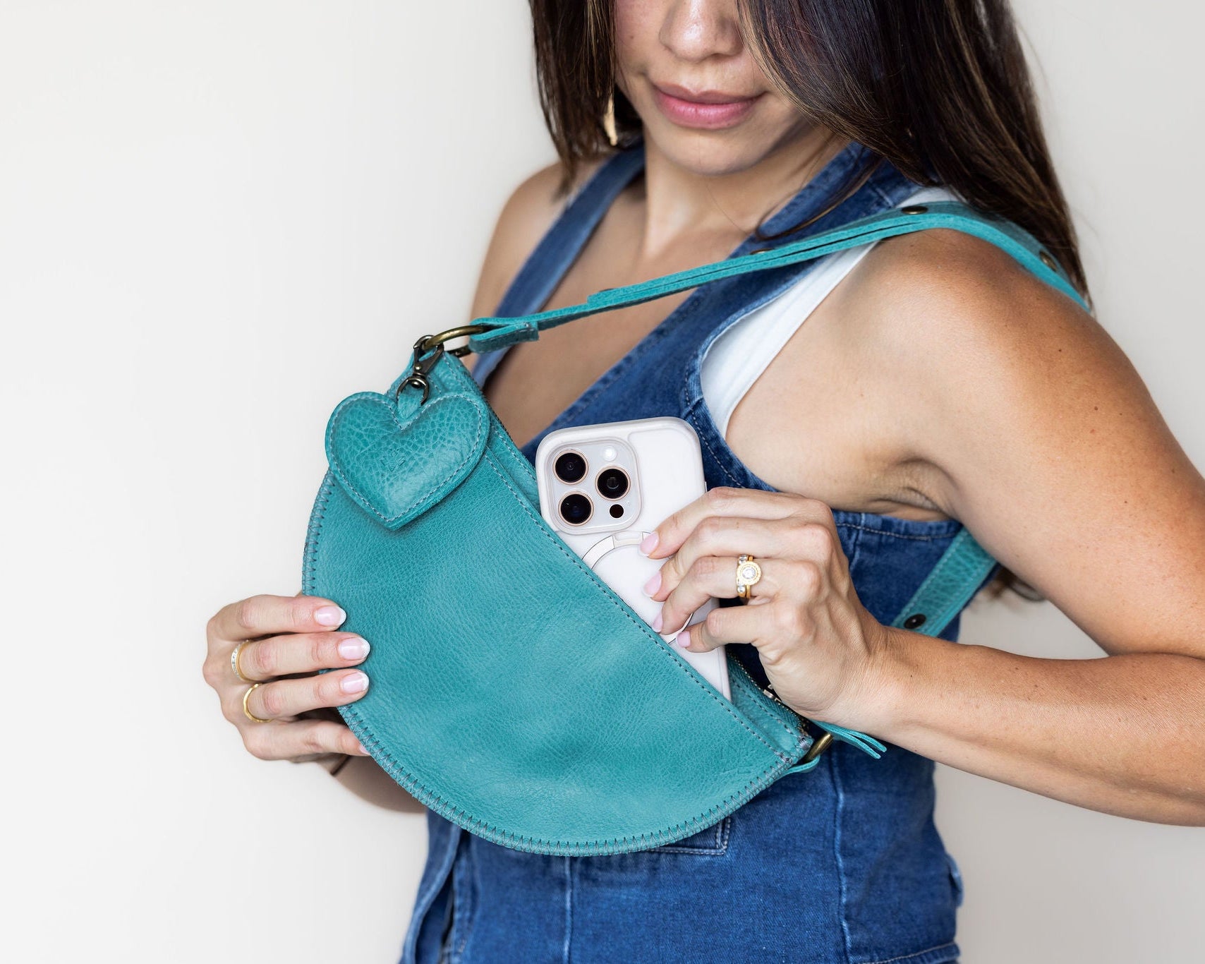 A woman wearing denim overalls holds a teal leather half-moon shoulder bag with a heart-shaped charm. A white smartphone is tucked into the bag's front pocket.