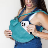 A woman wearing denim overalls holds a teal leather half-moon shoulder bag with a heart-shaped charm. A white smartphone is tucked into the bag's front pocket.