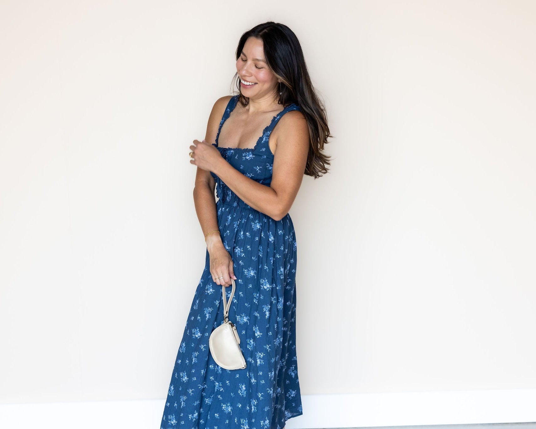 A woman wearing a blue floral print maxi dress and holding a small cream-colored handbag, posing against a plain white wall.